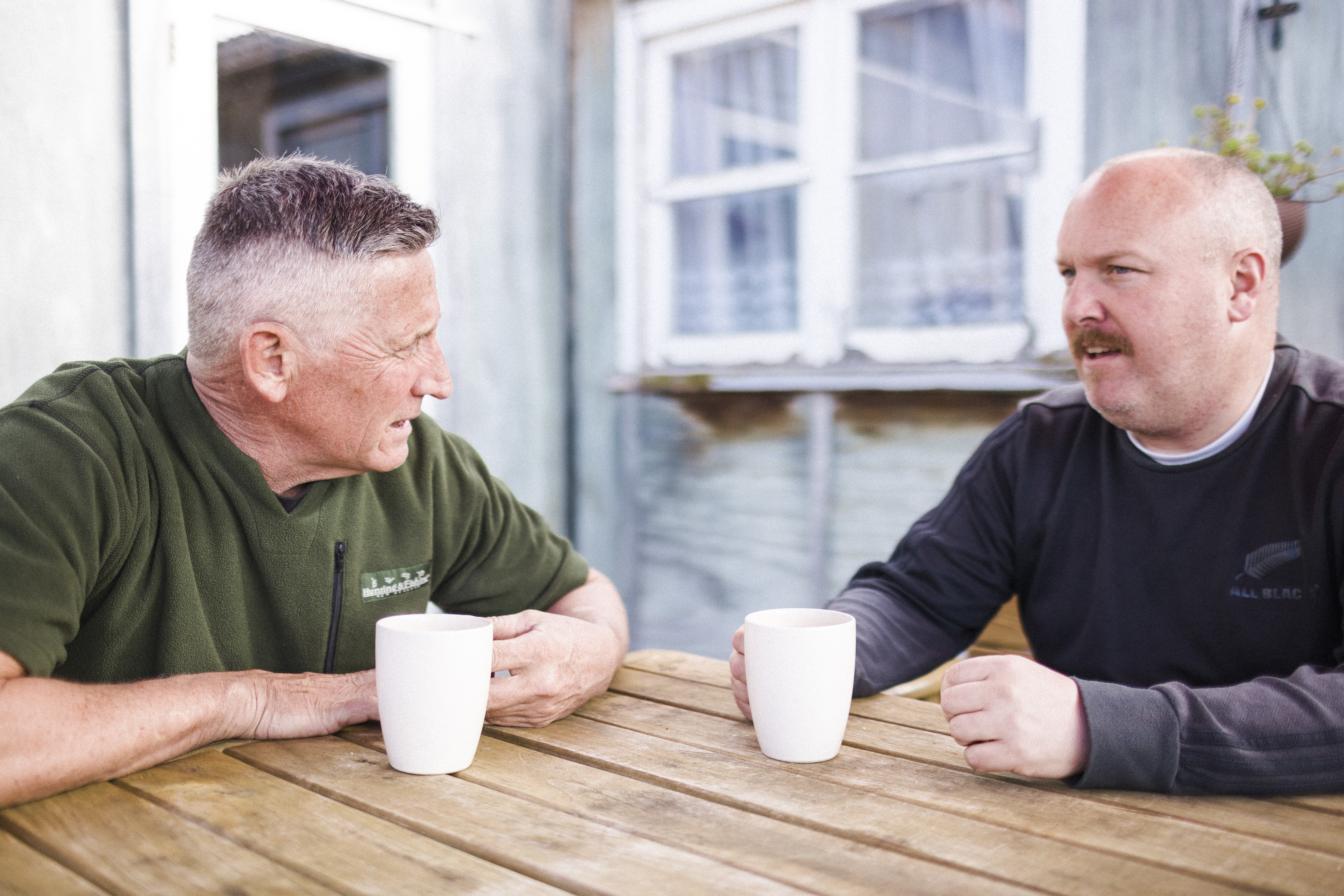 Two men sitting outside at a table talking with cups of tea