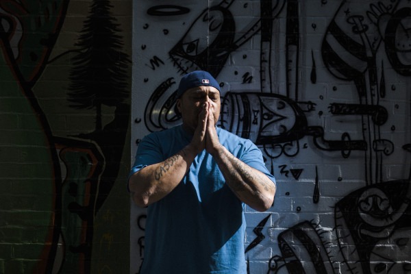 Man standing in front of wall with graffiti, eyes closed and hands in praying motion